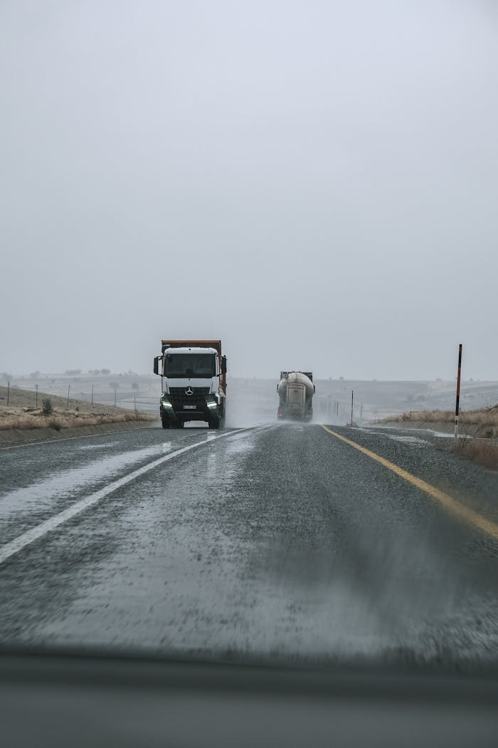 Two trucks driving on a rainy highway with overcast skies and empty roadsides.
